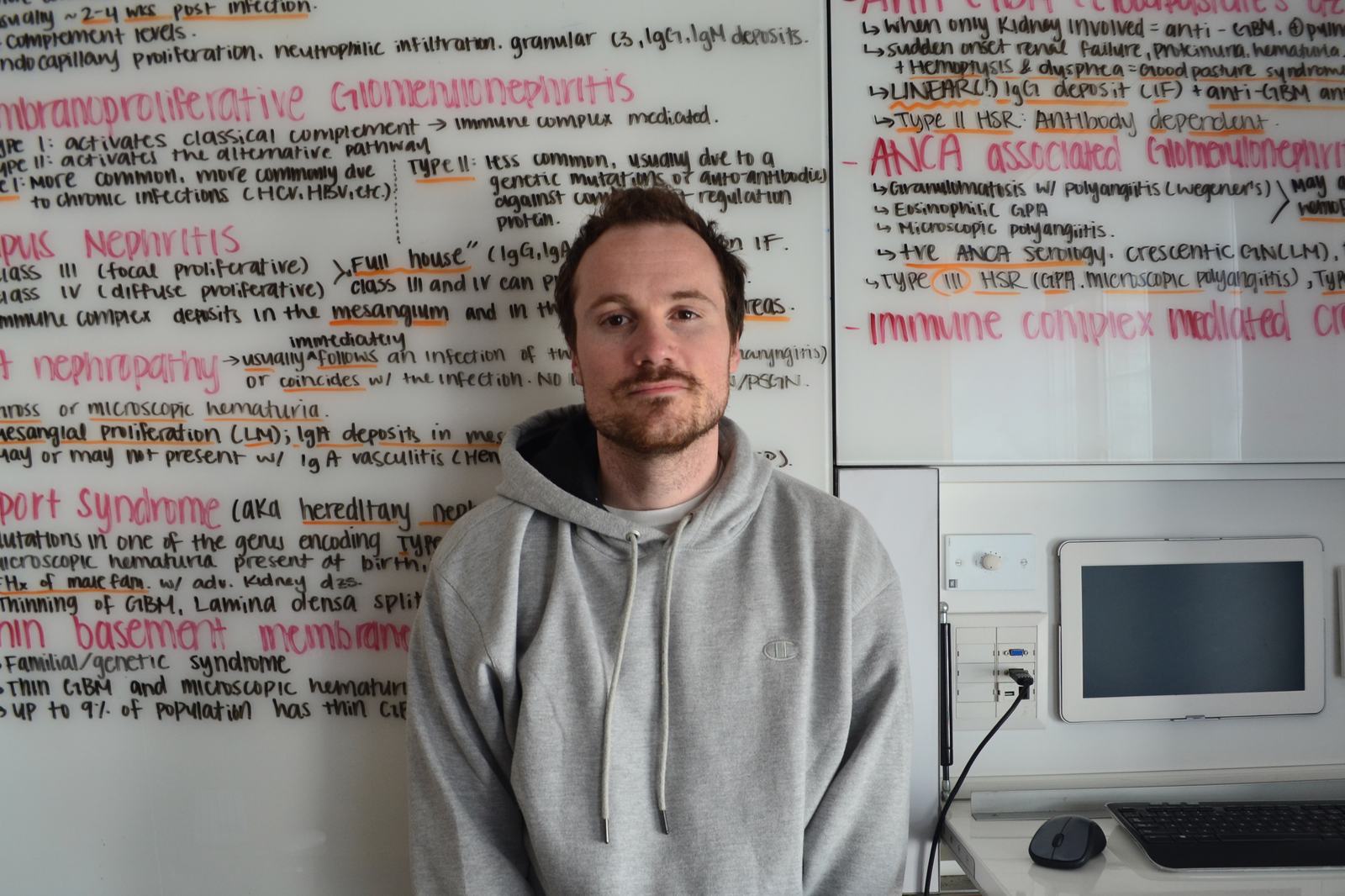 J Lind in a grey hoodie, standing in front of a whiteboard covered in handwritten medical notes