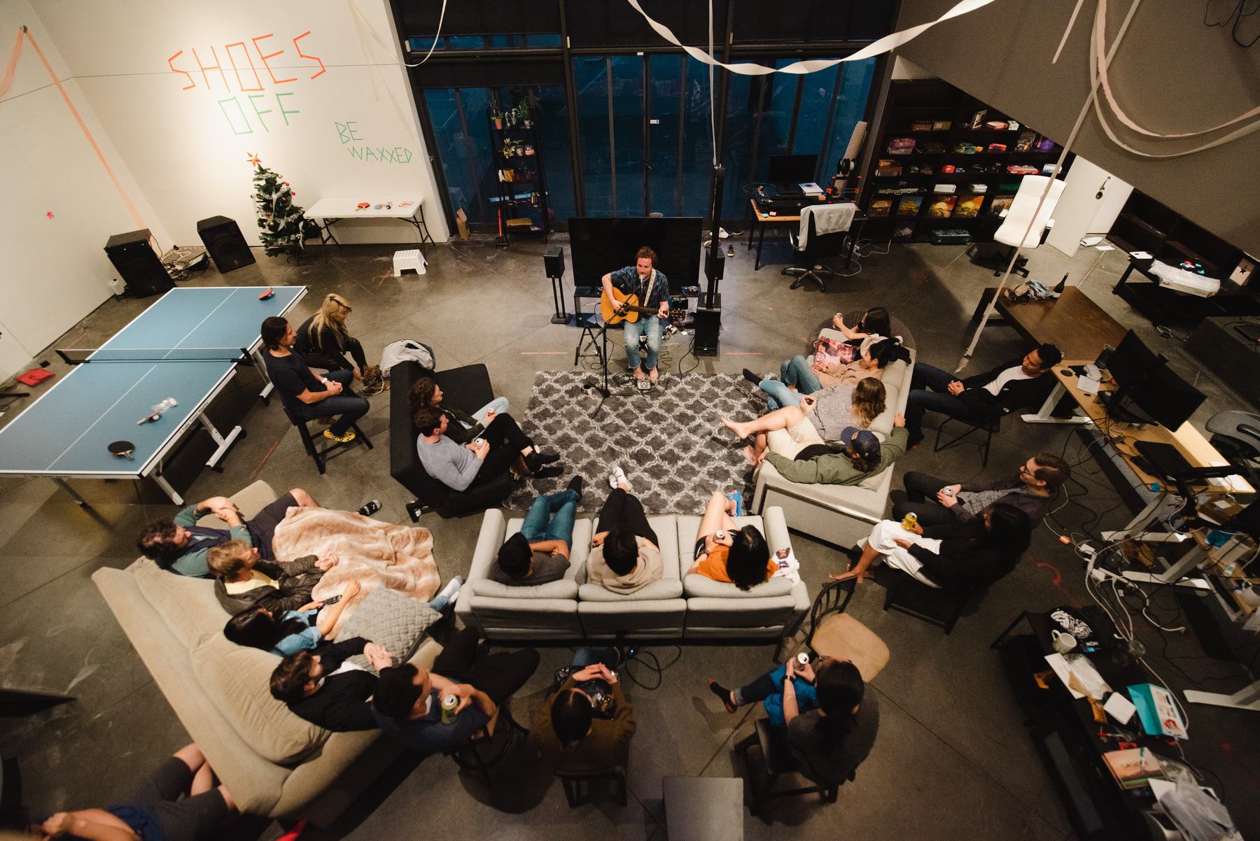Seen from above: J Lind playing guitar on a rug in a loft, with about twenty people listening from sofas and chairs around him