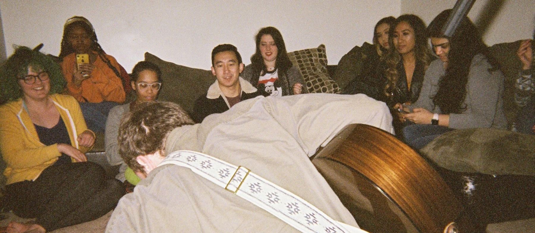 A living room crowd packed onto a sofa, smiling at J Lind as he bows over his guitar